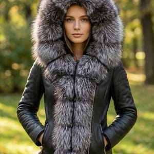 Puede incluir: Chaqueta de cuero negro con una gran capucha y cuello forrados de piel gris plateado. La chaqueta tiene cierre de cremallera y bolsillos. La modelo lleva la chaqueta al aire libre en un entorno natural.