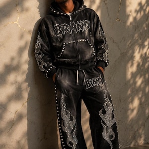 Puede incluir: Chándal de terciopelo negro con detalles de pedrería. La sudadera con cremallera y el pantalón a juego presentan la palabra "BRAND" en pedrería. La sudadera y el pantalón tienen diseños de llamas y están adornados con pedrería y tachuelas.