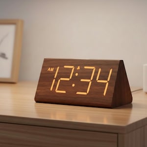 May include: A wooden digital alarm clock displaying "12:34 AM". The clock has a triangular shape and a warm, amber-colored digital display. The clock is made of wood with a visible wood grain.