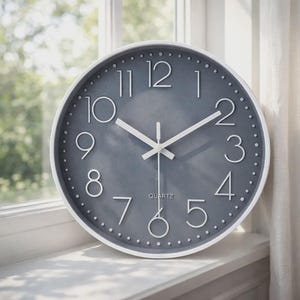 May include: A round, gray quartz clock with a white frame. The clock face features white numerals and hands, with the word "QUARTZ" at the bottom. The clock is set against a window with a blurred green background.