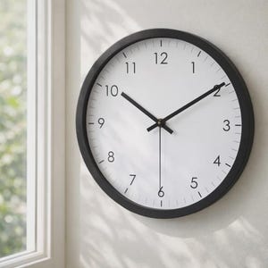 May include: A circular wall clock with a black surround and a white face. The clock features black hands and black numerals indicating the hours. It is mounted on a white wall, near a window.