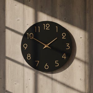 May include: A circular black wall clock with gold hands and numerals. The clock face is plain black, with the numbers in a modern font. The clock is mounted on a light-coloured wooden wall.