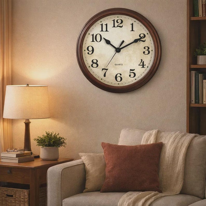 May include: A round wall clock with a dark brown wooden frame. The clock face is off-white with black numerals and hands. The clock is mounted on a beige wall, above a sofa with pillows and a throw blanket. The clock is a decorative home decor item.