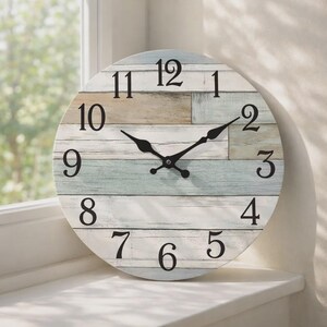 May include: Circular wall clock with a distressed wood-effect face. The clock face has a mix of white, light blue, and tan horizontal planks. Black numerals and hands provide contrast. The clock is displayed on a white windowsill.