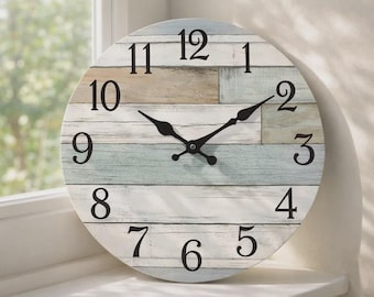 May include: Round wall clock with a distressed wood-look face. The clock face features a mix of white, light blue, and tan horizontal planks. Black numerals and hands provide contrast. The clock is displayed on a white windowsill.