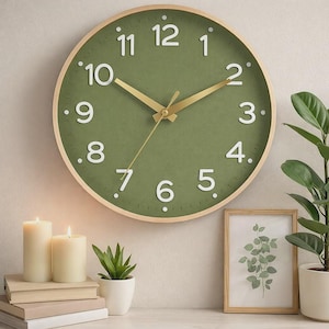May include: A round wall clock with a green face and white numerals. The clock has gold-colored hands and a light wood frame. The clock is surrounded by candles, plants, and a framed botanical print, creating a cozy, decorative scene.
