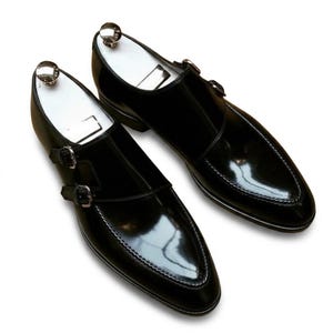 May include: A pair of black leather double monk strap shoes with a high-gloss finish, featuring silver-toned metal buckles and white shoe trees inserted inside each shoe to maintain their structured, pointed-toe shape and sleek profile.