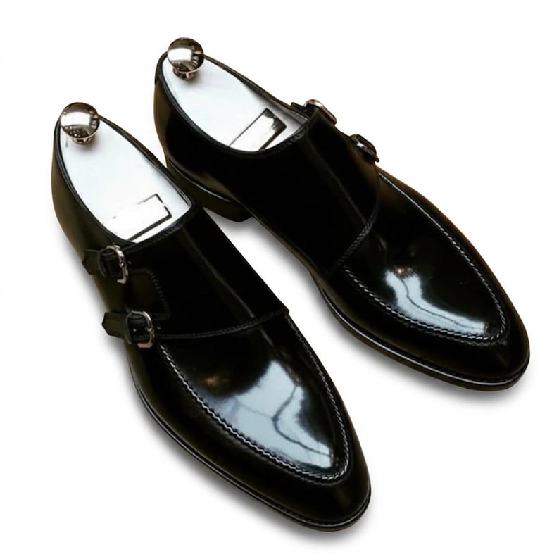 May include: A pair of black leather double monk strap shoes with a high-gloss finish, featuring silver-toned metal buckles and white shoe trees inserted inside each shoe to maintain their structured, pointed-toe shape and sleek profile.
