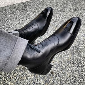 May include: A pair of black leather lace-up boots featuring a polished cap toe design and a stacked heel. The boots are worn with grey trousers that have a cuffed hem, showcasing the sleek silhouette and smooth texture of the black leather material.