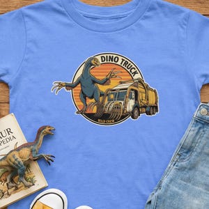 以下が含まれることがあります： 恐竜とゴミ収集車が描かれた円形グラフィックが特徴のライトブルーのTシャツ。「Dino Truck」と「Wild Crew」の文字。おもちゃのダンプトラック、恐竜のフィギュア、本も見える。