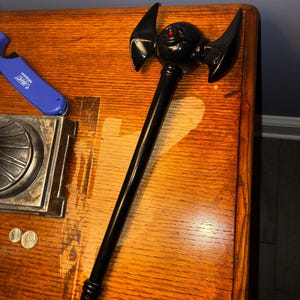 May include: A black, stylized axe with a rounded head and two curved wings. The head features a single red eye. The axe is placed on a wooden surface, with a blue lighter and coins visible in the background.