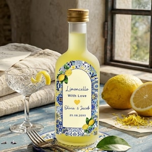 May include: A bottle of limoncello with a gold cap and a decorative label featuring lemons and blue floral patterns. The label reads "Limoncello With Love" and includes a couple's names and a date. A cocktail glass with a lemon twist sits nearby.