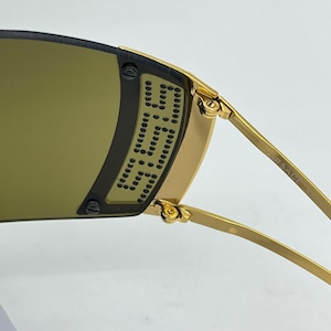 May include: Gold-tone sunglasses featuring a wrap-around dark lens with a perforated metal side shield detail. The temple arm is engraved with the text VERSACE. The frame includes visible screws securing the lens to the gold metal structure.
