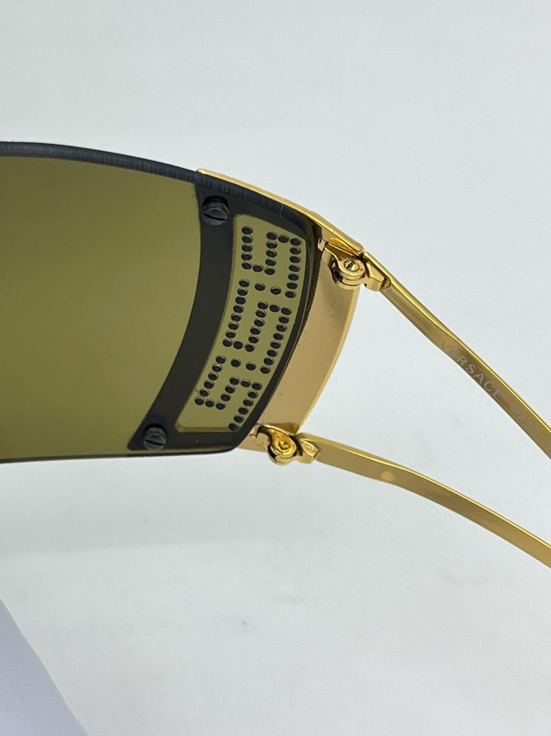 May include: Gold-tone sunglasses featuring a wrap-around dark lens with a perforated metal side shield detail. The temple arm is engraved with the text VERSACE. The frame includes visible screws securing the lens to the gold metal structure.