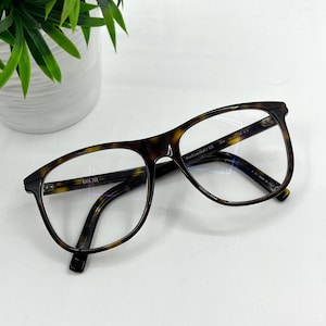 Op de afbeelding: Bril met schildpadpatroon en heldere glazen. De monturen zijn een mix van bruin en zwart, met het woord "DIOR" op de linker poot. De bril heeft een vierkante vorm.