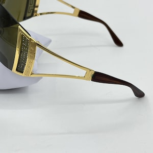 May include: Shield sunglasses featuring a gold-tone metal frame with a Greek key pattern and embedded clear crystals on the temples. The lenses are dark brown, and the temple tips are a dark brown tortoiseshell-style material with curved ends.