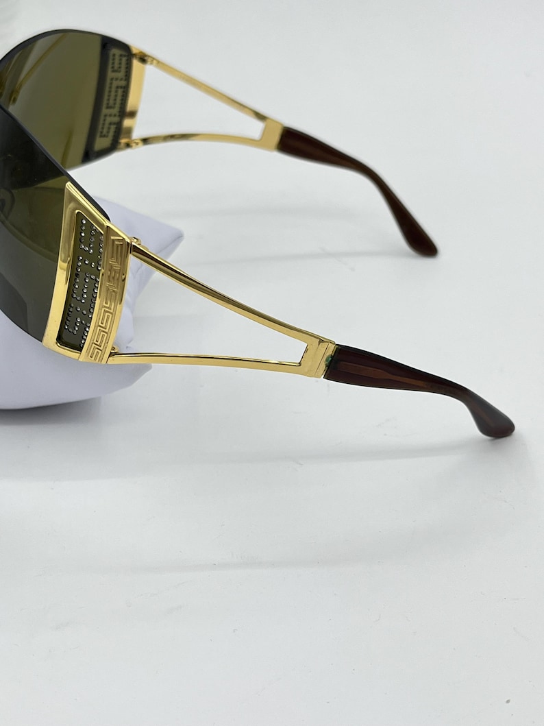 May include: Shield sunglasses featuring a gold-tone metal frame with a Greek key pattern and embedded clear crystals on the temples. The lenses are dark brown, and the temple tips are a dark brown tortoiseshell-style material with curved ends.