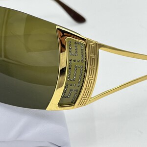 May include: Gold-tone sunglasses featuring dark olive-tinted lenses. The frame includes a decorative gold panel on the temple adorned with small clear crystals and a geometric Greek key pattern. The temple arms are thin and gold-toned.