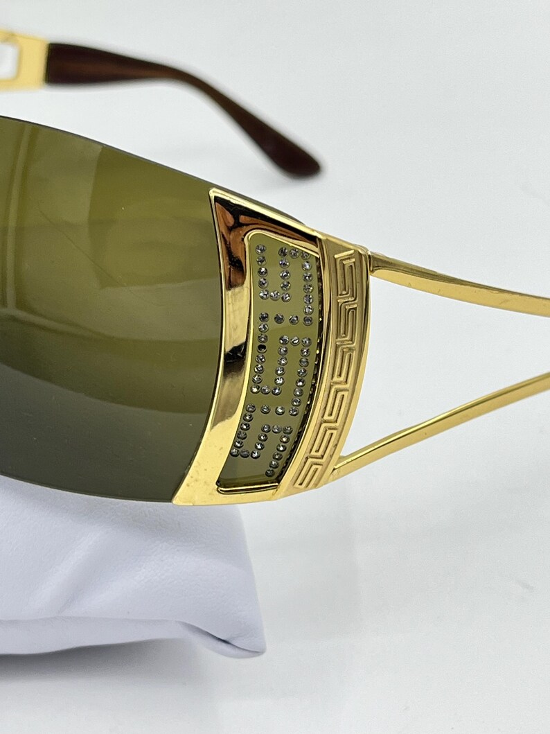 May include: Gold-tone sunglasses featuring dark olive-tinted lenses. The frame includes a decorative gold panel on the temple adorned with small clear crystals and a geometric Greek key pattern. The temple arms are thin and gold-toned.
