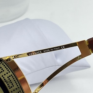 May include: Gold-toned metal sunglasses temple arm featuring the text VERSACE MADE IN ITALY CE printed in black. The frame includes a decorative side panel with a repeating circular pattern and a dark brown temple tip.