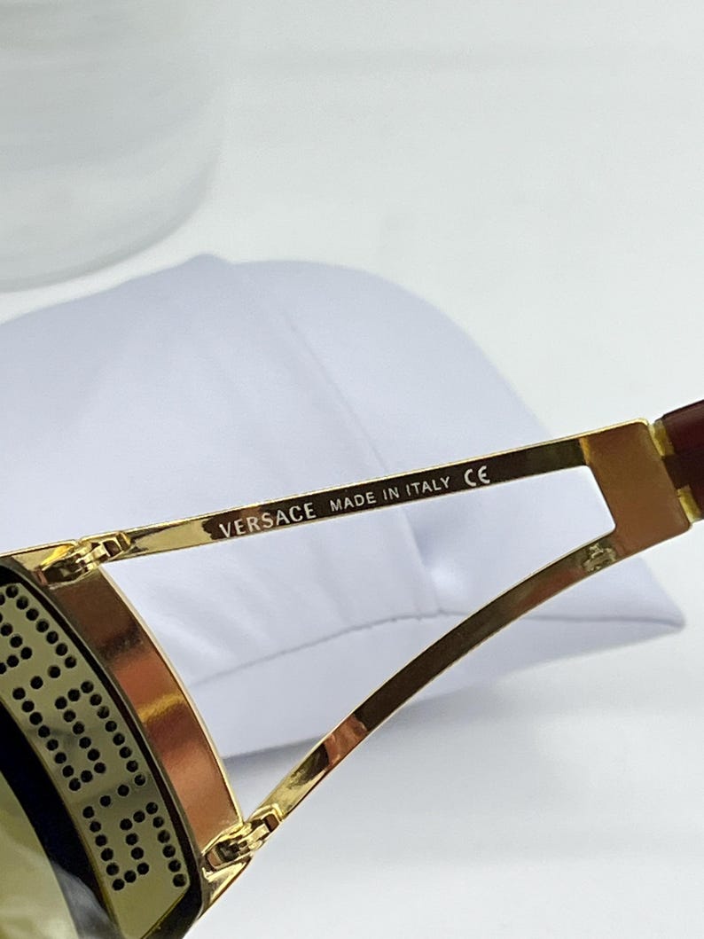 May include: Gold-toned metal sunglasses temple arm featuring the text VERSACE MADE IN ITALY CE printed in black. The frame includes a decorative side panel with a repeating circular pattern and a dark brown temple tip.