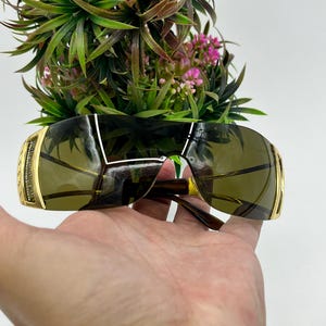 May include: Shield sunglasses featuring a single continuous dark-tinted lens and gold-tone metal temples. The side hinges are embellished with small clear crystals set in a gold frame, providing a metallic finish to the wrap-around eyewear design.