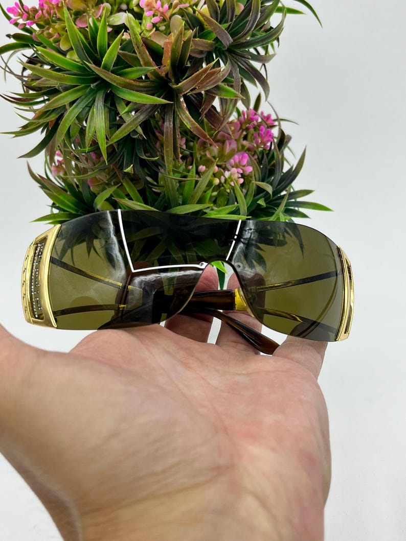 May include: Shield sunglasses featuring a single continuous dark-tinted lens and gold-tone metal temples. The side hinges are embellished with small clear crystals set in a gold frame, providing a metallic finish to the wrap-around eyewear design.