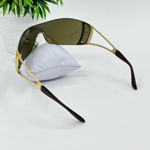 May include: Shield sunglasses featuring a single continuous dark brown lens and a gold-tone metal frame. The temples are thin gold metal with dark brown tips. A decorative gold-tone rectangular accent with a Greek key pattern is visible on the side.
