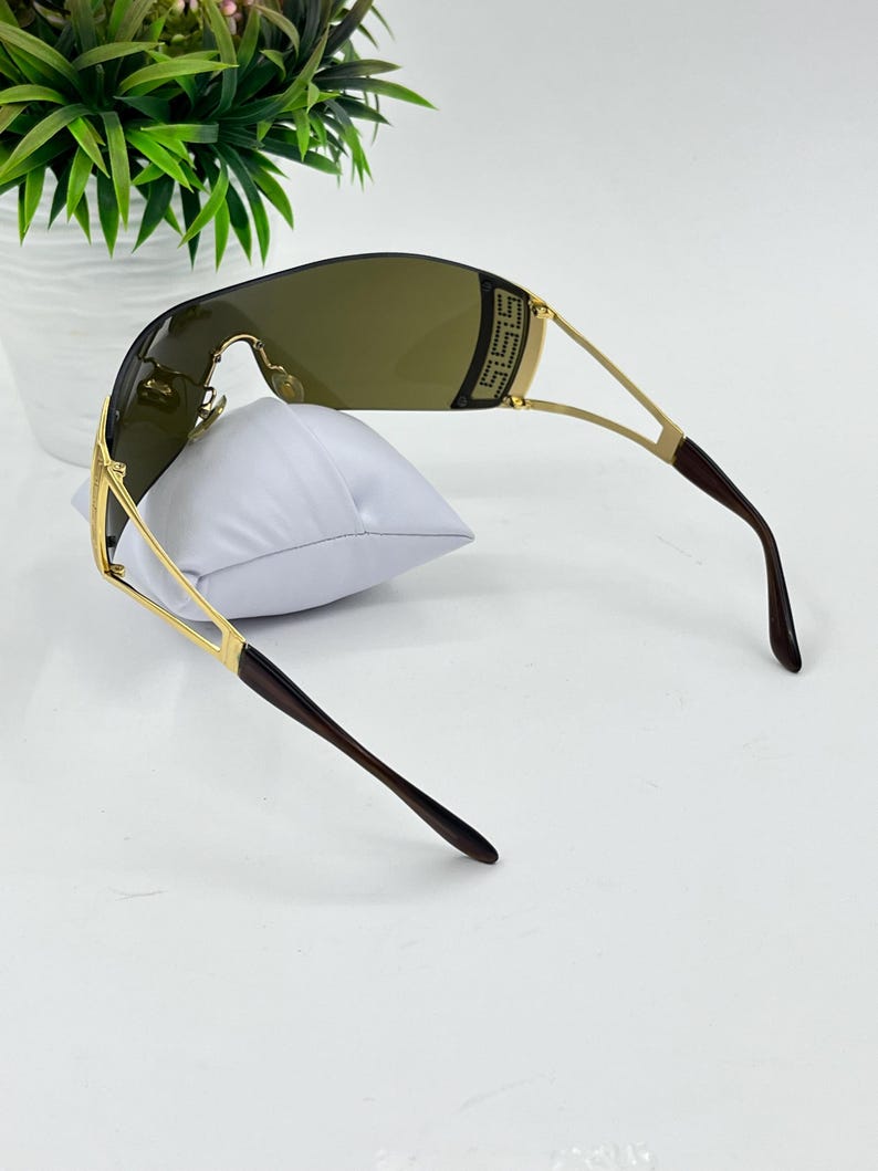 May include: Shield sunglasses featuring a single continuous dark brown lens and a gold-tone metal frame. The temples are thin gold metal with dark brown tips. A decorative gold-tone rectangular accent with a Greek key pattern is visible on the side.
