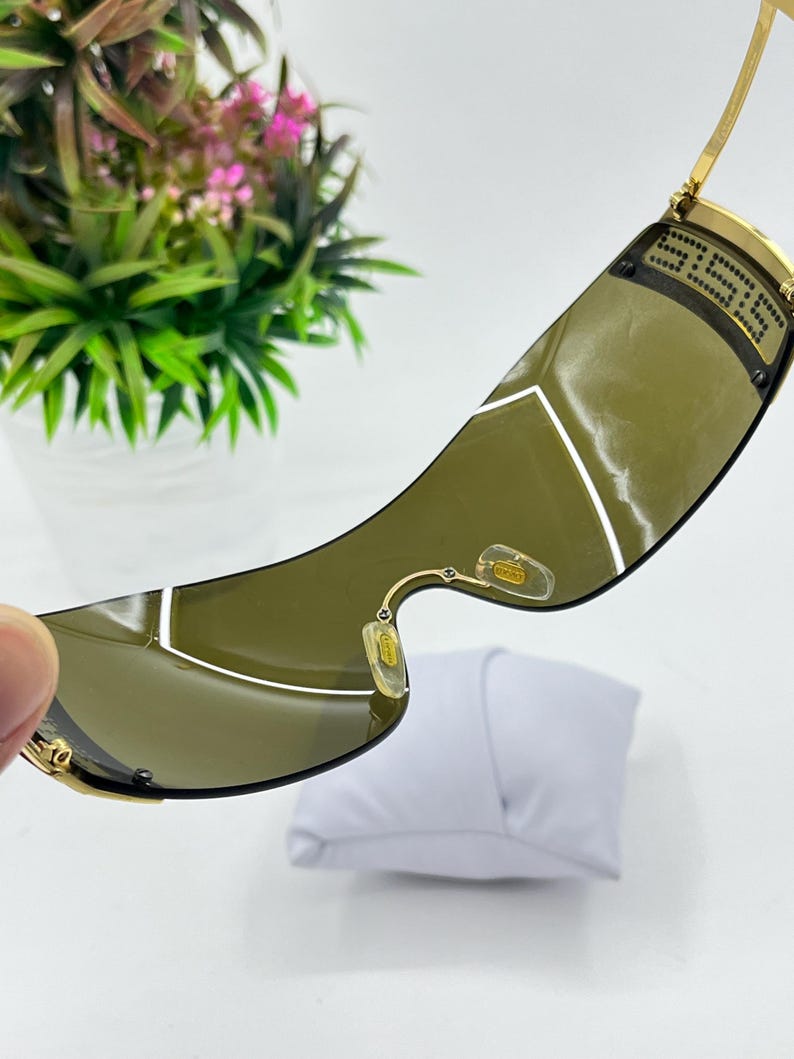 May include: Gold-tone sunglasses featuring a single-piece wrap-around dark olive lens. The frame includes thin gold metal temples with a black geometric pattern near the hinges and clear nose pads with gold-tone branding visible on the interior.