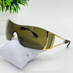 May include: Shield-style sunglasses featuring a single continuous dark olive-tinted lens. The frame is gold-toned metal with a decorative Greek key pattern on the temples and a textured panel embedded with small clear crystals on the side hinges.