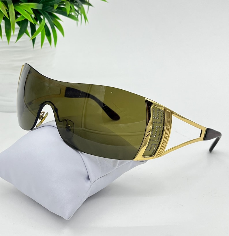 May include: Shield-style sunglasses featuring a single continuous dark olive-tinted lens. The frame is gold-toned metal with a decorative Greek key pattern on the temples and a textured panel embedded with small clear crystals on the side hinges.