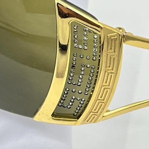 May include: Gold-tone sunglasses featuring a wide, polished frame with a Greek key pattern on the temple. The side panel is inlaid with small, clear crystals arranged in a grid pattern, paired with dark, olive-tinted lenses for a sleek look.