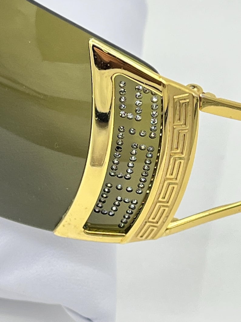 May include: Gold-tone sunglasses featuring a wide, polished frame with a Greek key pattern on the temple. The side panel is inlaid with small, clear crystals arranged in a grid pattern, paired with dark, olive-tinted lenses for a sleek look.