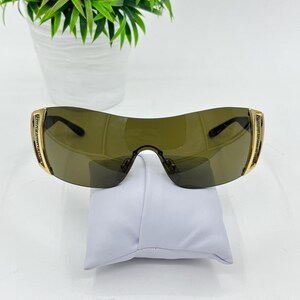 May include: Shield-style sunglasses featuring a single continuous dark olive-tinted lens, gold-tone metal frames, and side temples embellished with rows of small clear crystals. The design includes a bridge with adjustable nose pads.