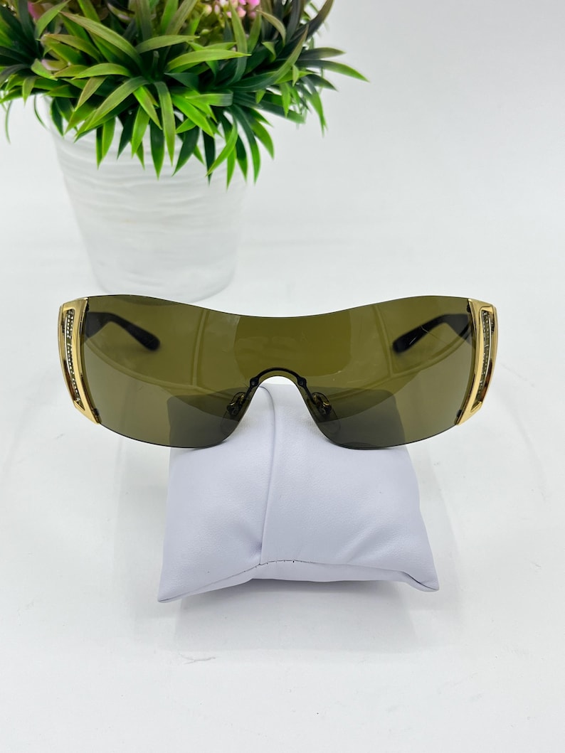 May include: Shield-style sunglasses featuring a single continuous dark olive-tinted lens, gold-tone metal frames, and side temples embellished with rows of small clear crystals. The design includes a bridge with adjustable nose pads.