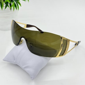 May include: Shield sunglasses featuring a single continuous dark olive green lens and gold-tone metal temples. The side hinges are embellished with a decorative Greek key pattern and small embedded crystals, connected to thin gold-tone metal arms.