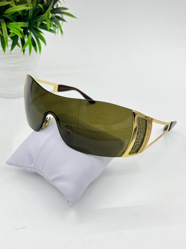 May include: Shield sunglasses featuring a single continuous dark olive green lens and gold-tone metal temples. The side hinges are embellished with a decorative Greek key pattern and small embedded crystals, connected to thin gold-tone metal arms.