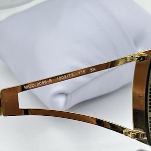 May include: Gold-toned metal sunglasses temple arm featuring the printed text MOD. 2058-B 1002/73 115 3N on the inner side. The frame arm has a split design near the hinge and a polished metallic finish, showing clear technical specifications.