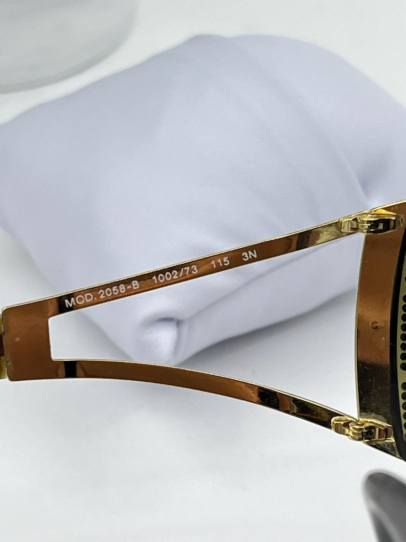 May include: Gold-toned metal sunglasses temple arm featuring the printed text MOD. 2058-B 1002/73 115 3N on the inner side. The frame arm has a split design near the hinge and a polished metallic finish, showing clear technical specifications.