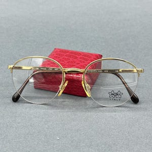 May include: Gold-tone semi-rimless spectacles with round lenses. The bridge is engraved with 135 FRAME ITALY GEP. The right lens features the Luxottica Italy logo. The temples have dark brown tips. The frames are positioned in front of a red case.