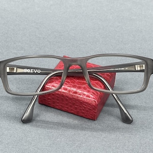 May include: Rectangular spectacles with a matte charcoal grey frame. The inner left temple features the white text "servo" and the inner right temple includes additional white text. The glasses are resting on a small, red, textured box.