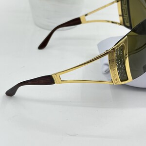 May include: Sunglasses with a gold-tone metal frame featuring a Greek key pattern on the temples. The lenses are dark-tinted and wrap around the sides. The temple tips are a dark brown tortoiseshell pattern, and the frame has a polished finish.