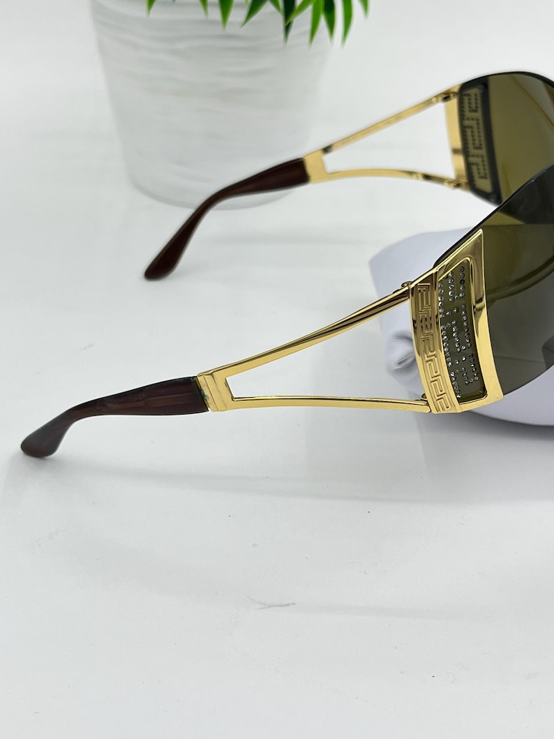 May include: Sunglasses with a gold-tone metal frame featuring a Greek key pattern on the temples. The lenses are dark-tinted and wrap around the sides. The temple tips are a dark brown tortoiseshell pattern, and the frame has a polished finish.