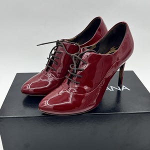 Puede incluir: Un par de zapatos Oxford de tacón alto de charol burdeos con cordones negros. Los zapatos están sobre una caja negra con texto blanco. Los tacones son delgados y los zapatos tienen un acabado brillante.