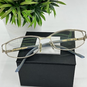 May include: Silver-framed spectacles with clear lenses and light blue temples. The glasses are displayed on a black box, with a green plant in the background. The frame has a rectangular shape.
