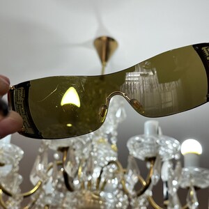 May include: Shield-style sunglasses featuring a single continuous amber-tinted lens and black frames. The temples are decorated with a gold-toned geometric pattern and small embedded crystals. The lens surface shows visible light surface scratches.