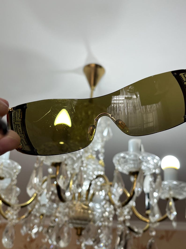 May include: Shield-style sunglasses featuring a single continuous amber-tinted lens and black frames. The temples are decorated with a gold-toned geometric pattern and small embedded crystals. The lens surface shows visible light surface scratches.