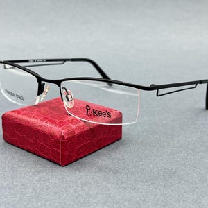 May include: Black semi-rimless eyeglasses with thin metal temples and a rectangular frame shape. The lenses feature the text L.Kee's and STAINLESS STEEL printed on them. The glasses are resting on a small, textured, bright red square box.