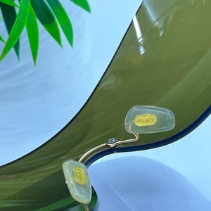 May include: A pair of olive green tinted sunglasses featuring a single continuous lens design. The nose pads are clear and translucent, with the word VERSACE embossed in gold lettering on the right nose pad, attached by a thin gold metal bridge.